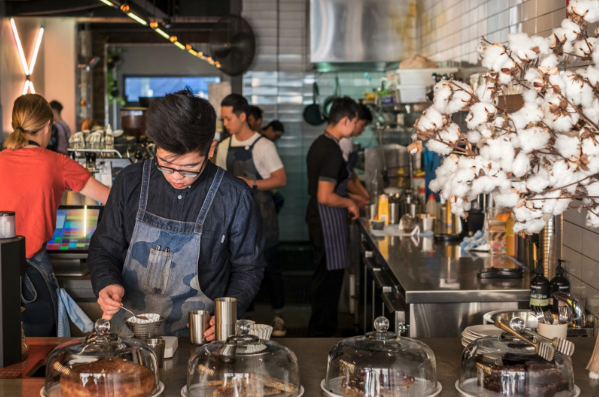 An animated café with several team members preparing orders, while a team member takes orders on an ipad via the cash register software.