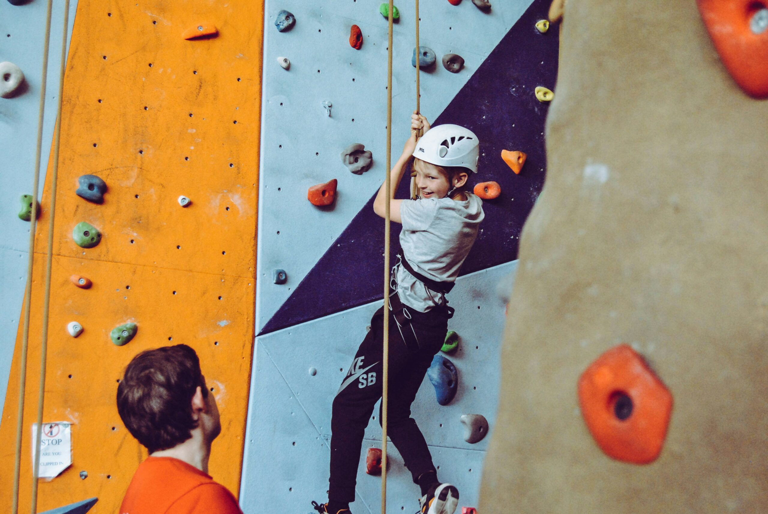 Photo of an indoor climbing gym. A climbing instructor helps a child climb the wall of holds.