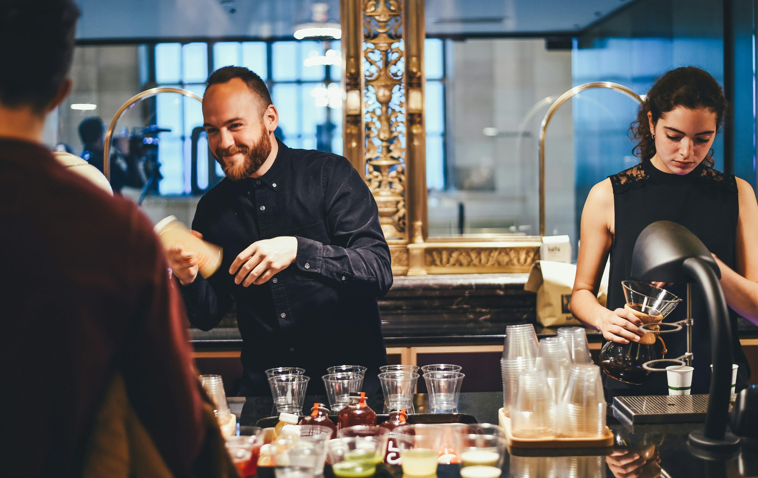 A waiter and waitress prepare drinks behind the bar. The waiter smiles at the customers.