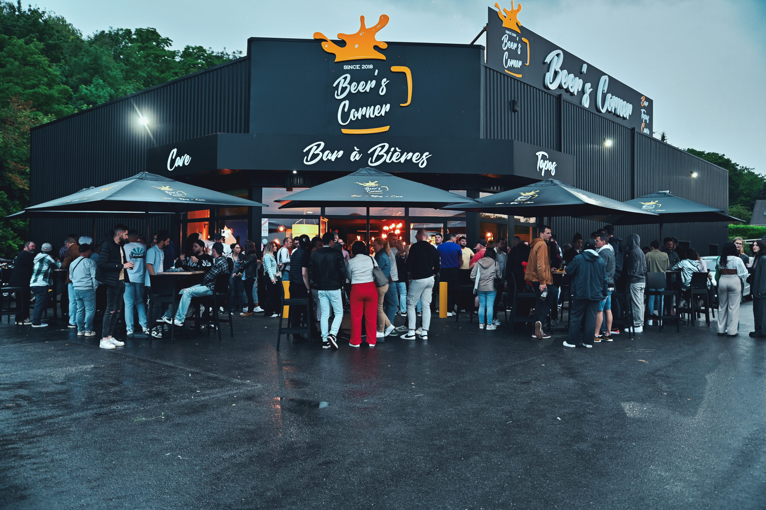 Exterior facade of a Beer's Corner bar, with a crowd of customers gathered under umbrellas, enjoying the convivial evening atmosphere. The facade is modern, with the sign visible.