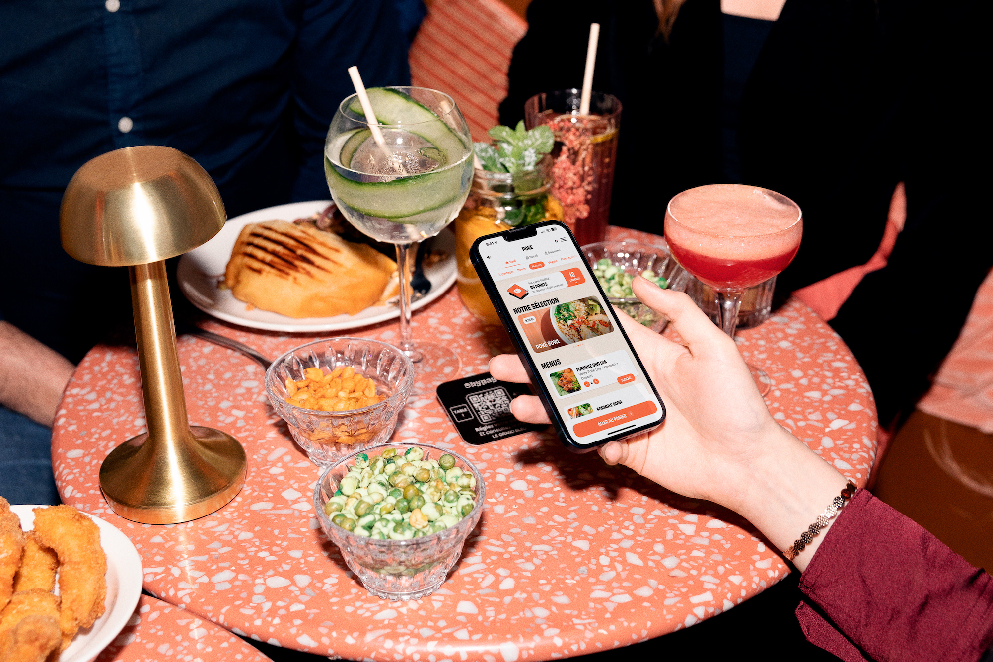 In a restaurant, a customer scans a QR code with her phone to consult the menu and order dishes. The QR code is placed in the center of the table, surrounded by several dishes and cocktails.