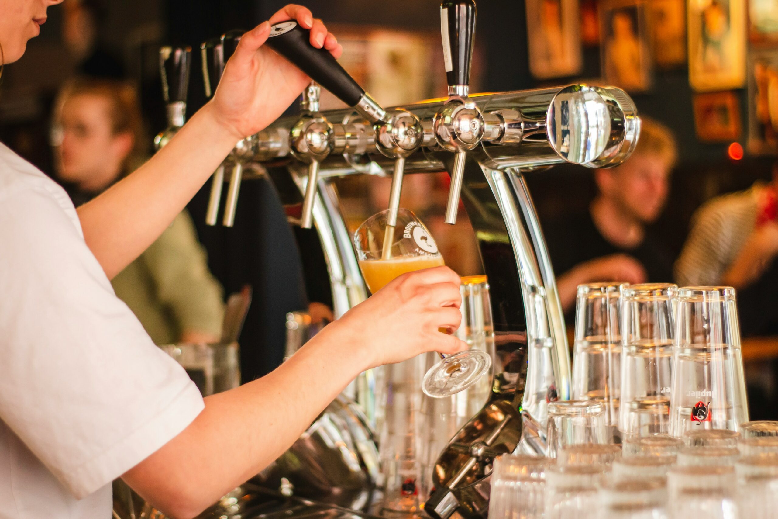 Cameriera che riempie un bicchiere di birra alla spina in un bar, con clienti sullo sfondo sfocato. La scena illustra l'attività di un locale di ristorazione.