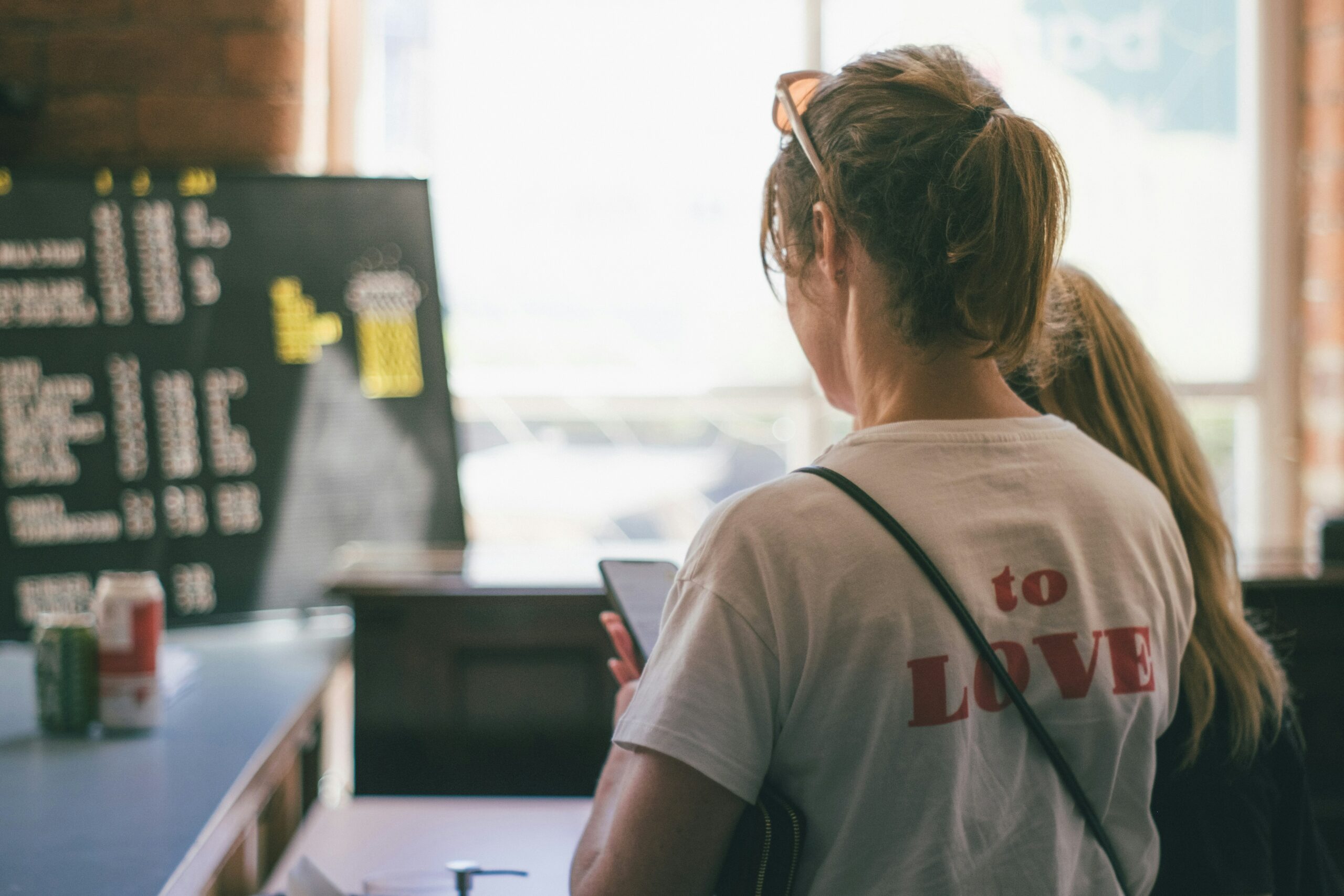 Cette photo montre une personne récupérant une commande en Click & Collect dans un restaurant. La femme, vue de dos, se tient devant un comptoir. Téléphone en main, elle est probablement en train de vérifier les informations de sa commande.