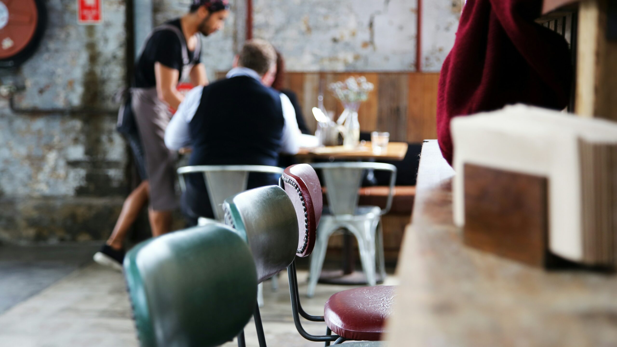 Cameriere che porta un piatto ai clienti seduti in un ristorante dall'arredamento industriale, con sedie di metallo in primo piano e un'atmosfera rilassata.