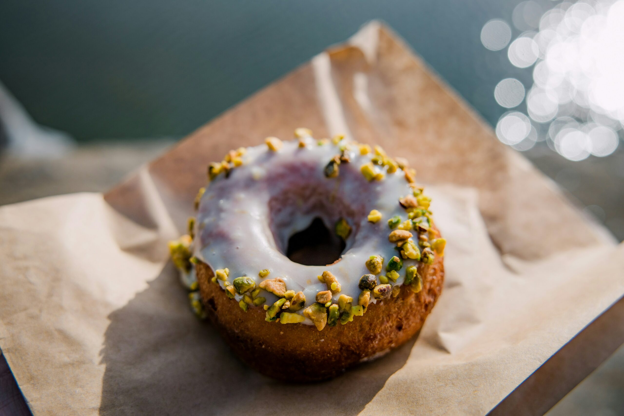 La photo présente un donut glacé, posé sur un morceau de papier brun, utilisé comme emballage. La photo a été prise en extérieur, l'arrière-plan flou montre un environnement extérieur lumineux.