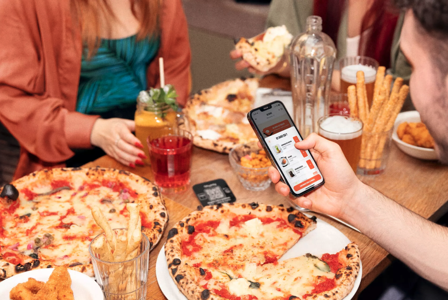 La photo montre des personnes assises au restaurant avec différents plats posés sur la table : des pizzas et des finger food. Une personne, de dos, tient un téléphone portable dans sa main et passe une commande dans le restaurant via le menu digital.