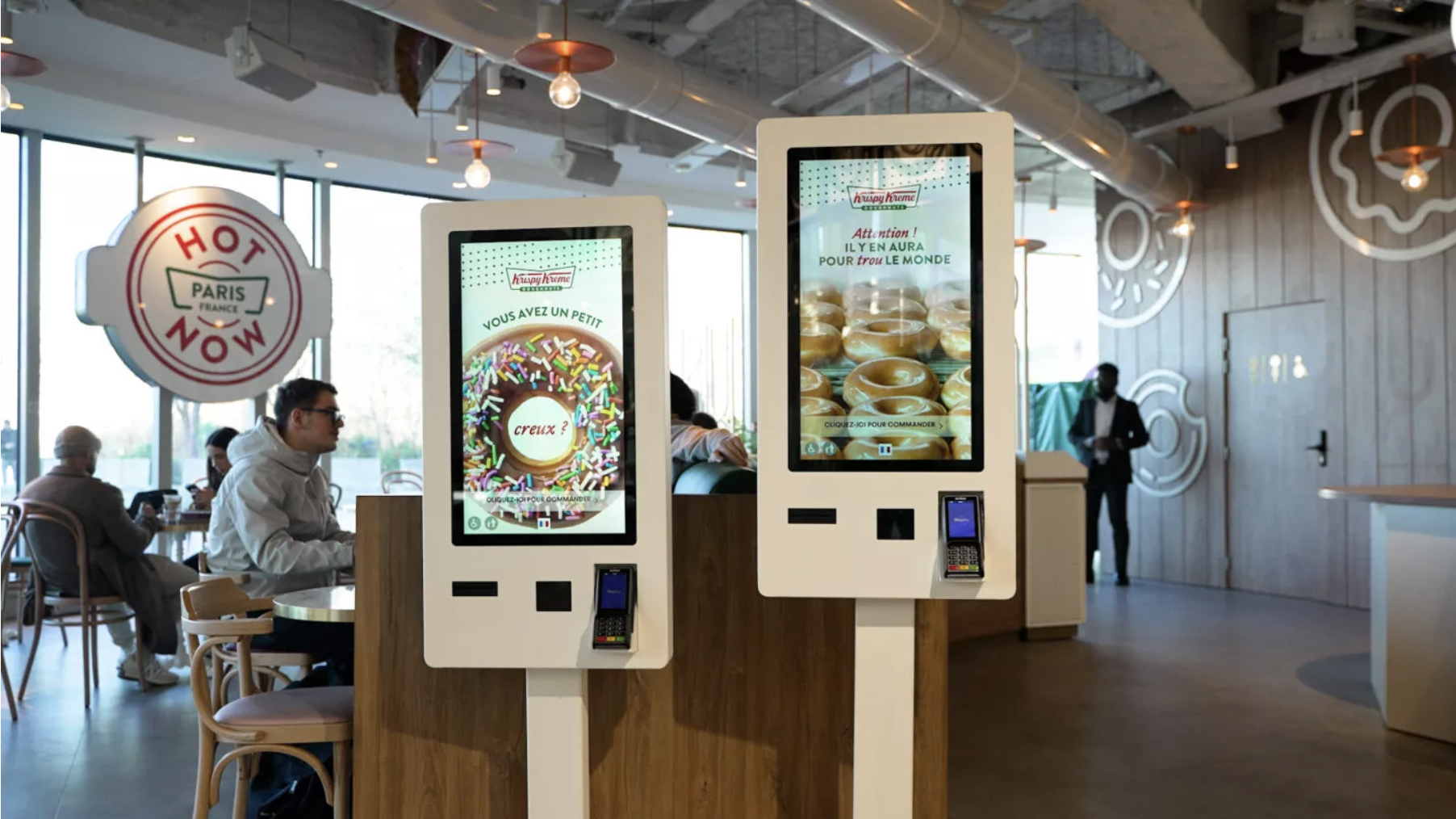 L’image montre deux bornes de commande sur pied Obypay installées dans la chaîne Krispy Kreme. Des écrans promotionnels affichent les dernières nouveautés de la marque.