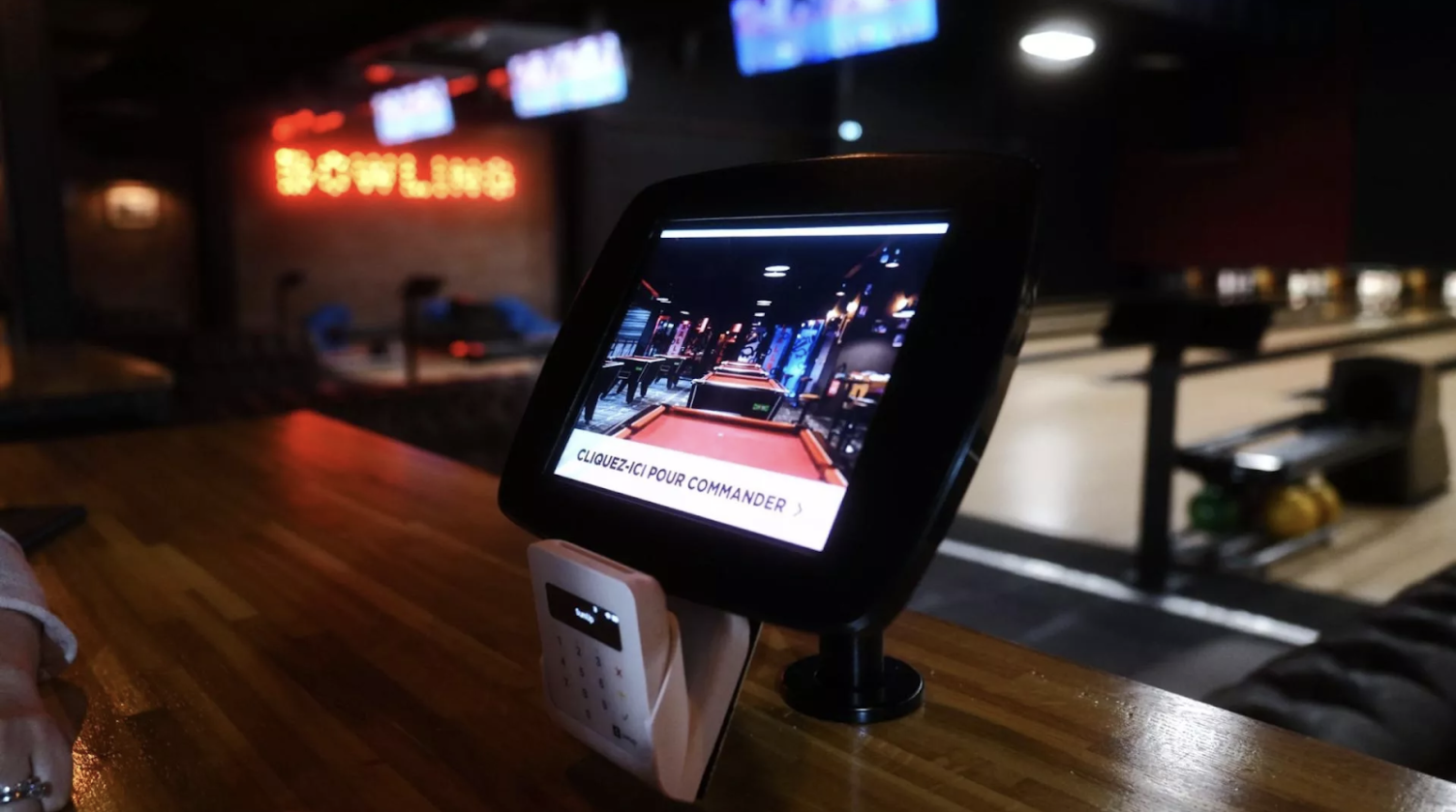 La photo montre une une salle de bowling. Au premier plan, une tablette de commandes permet aux clients de commander des snacks et des boissons directement depuis la piste de bowling. En arrière-plan, des pistes de bowling sont éclairées.