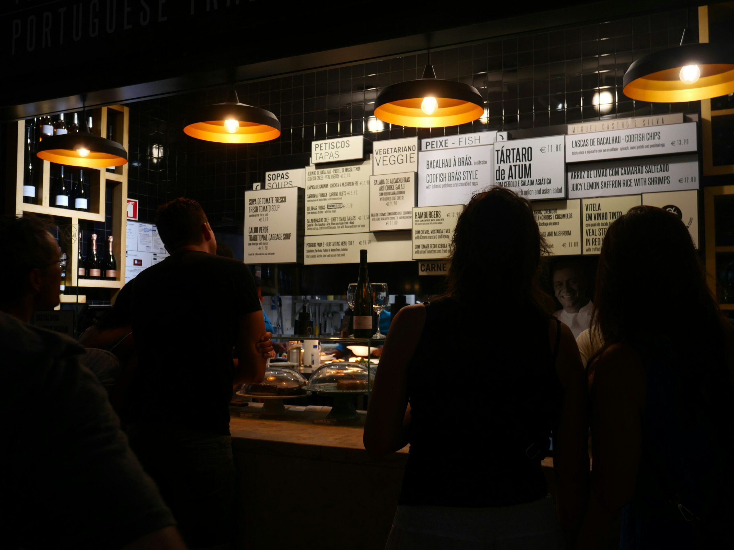 La photo montre un stand dans un food court, où des clients passent commande devant un comptoir. Sous la lumière tamisée des lampes, le menu propose des tapas, poissons, et plats végétariens.