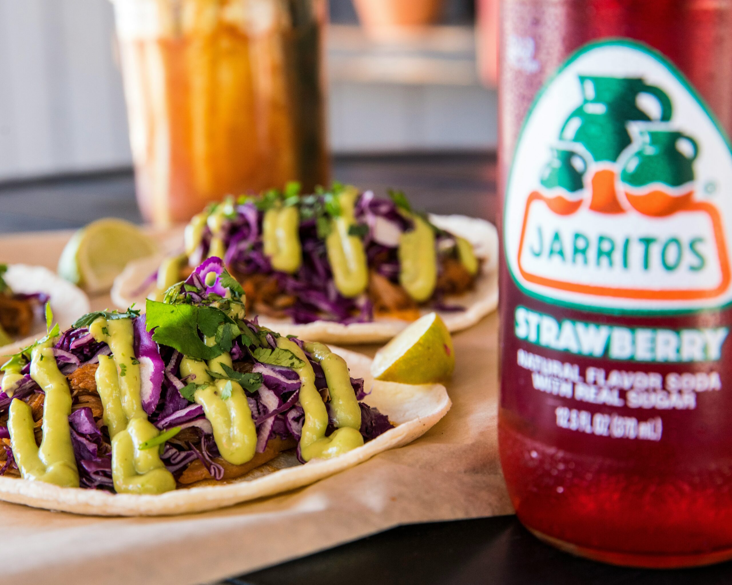 La photo montre des tacos garnis de chou violet et sauce, accompagnés d'une bouteille de soda Jarritos à la fraise.