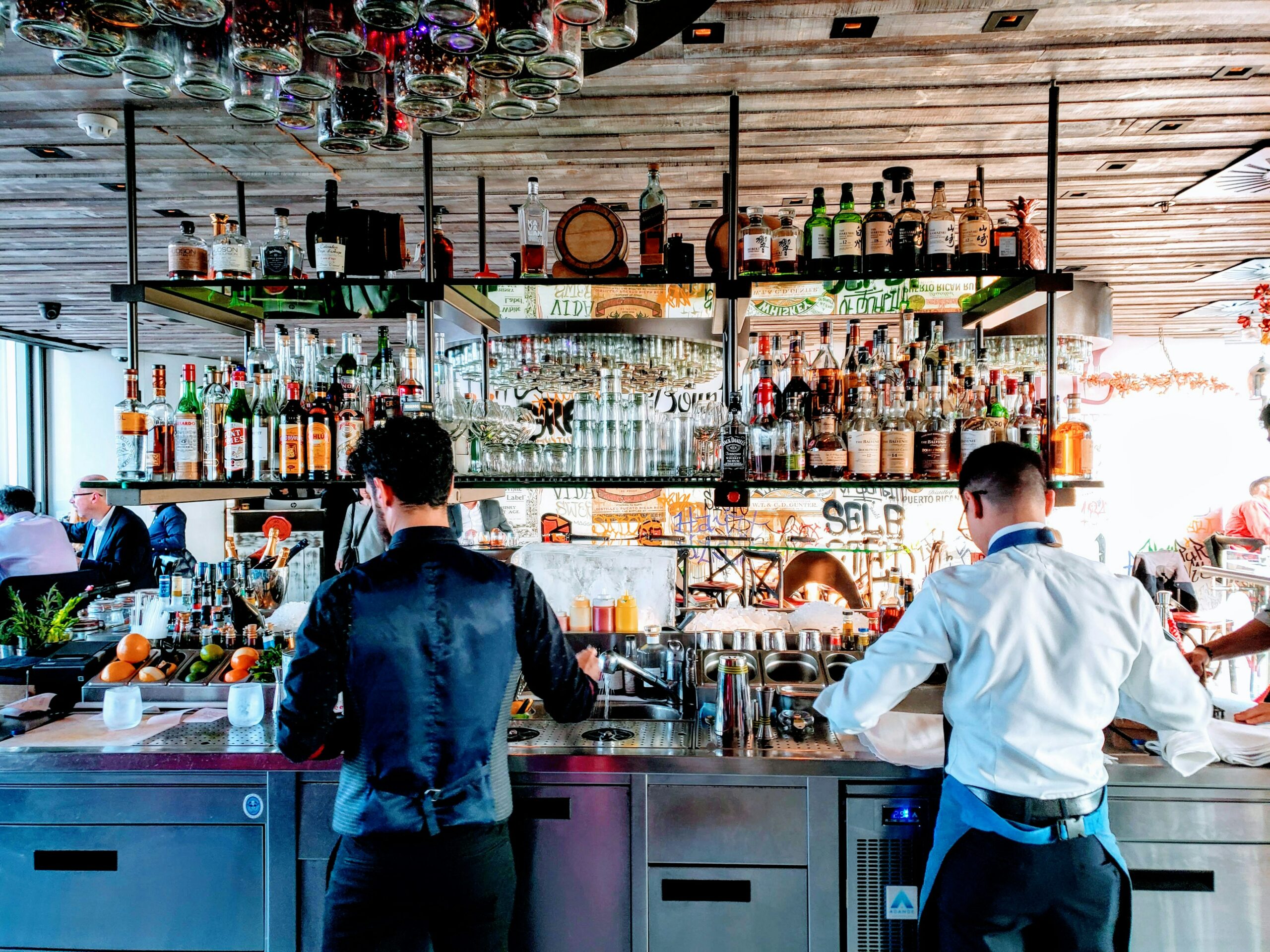 La photo montre deux barmans préparent des boissons derrière un bar, avec des étagères remplies de bouteilles d'alcool et de verres.