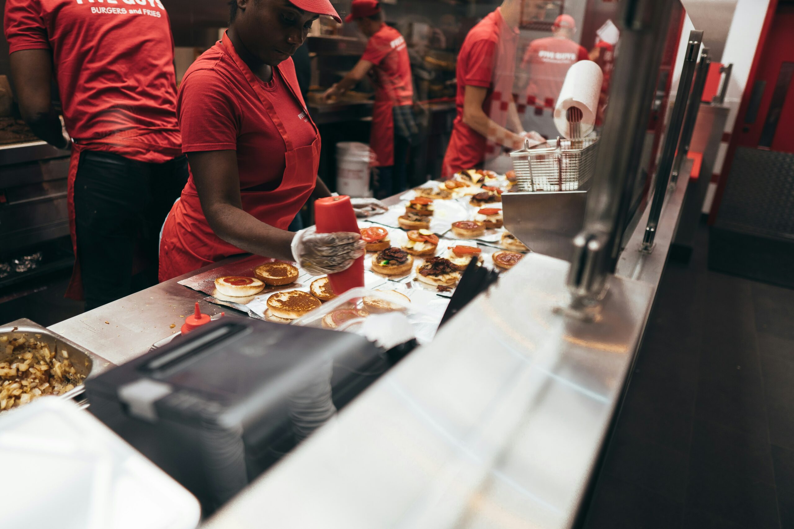 La photo montre des employés de restauration rapide, vêtus de rouge, qui préparent des hamburgers sur un comptoir de cuisine.