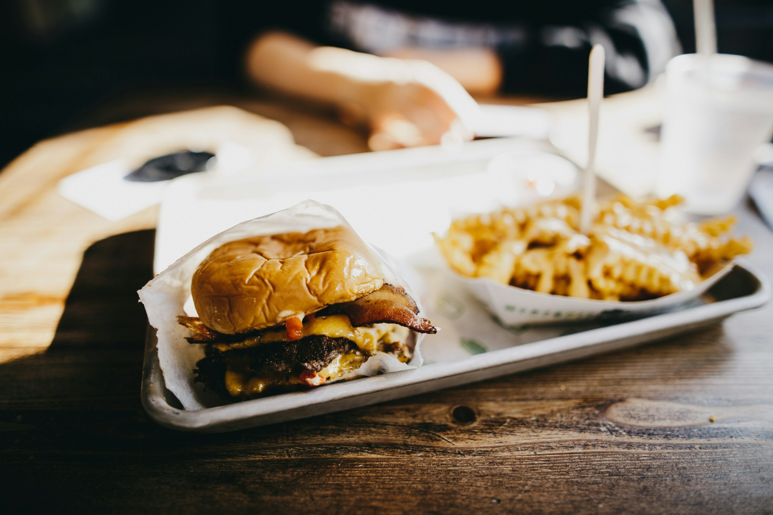 La photo montre un plateau de repas contenant un hamburger et des frites garnies de fromage. Le plateau est posé sur une table en bois devant une personne floue, en arrière-plan.