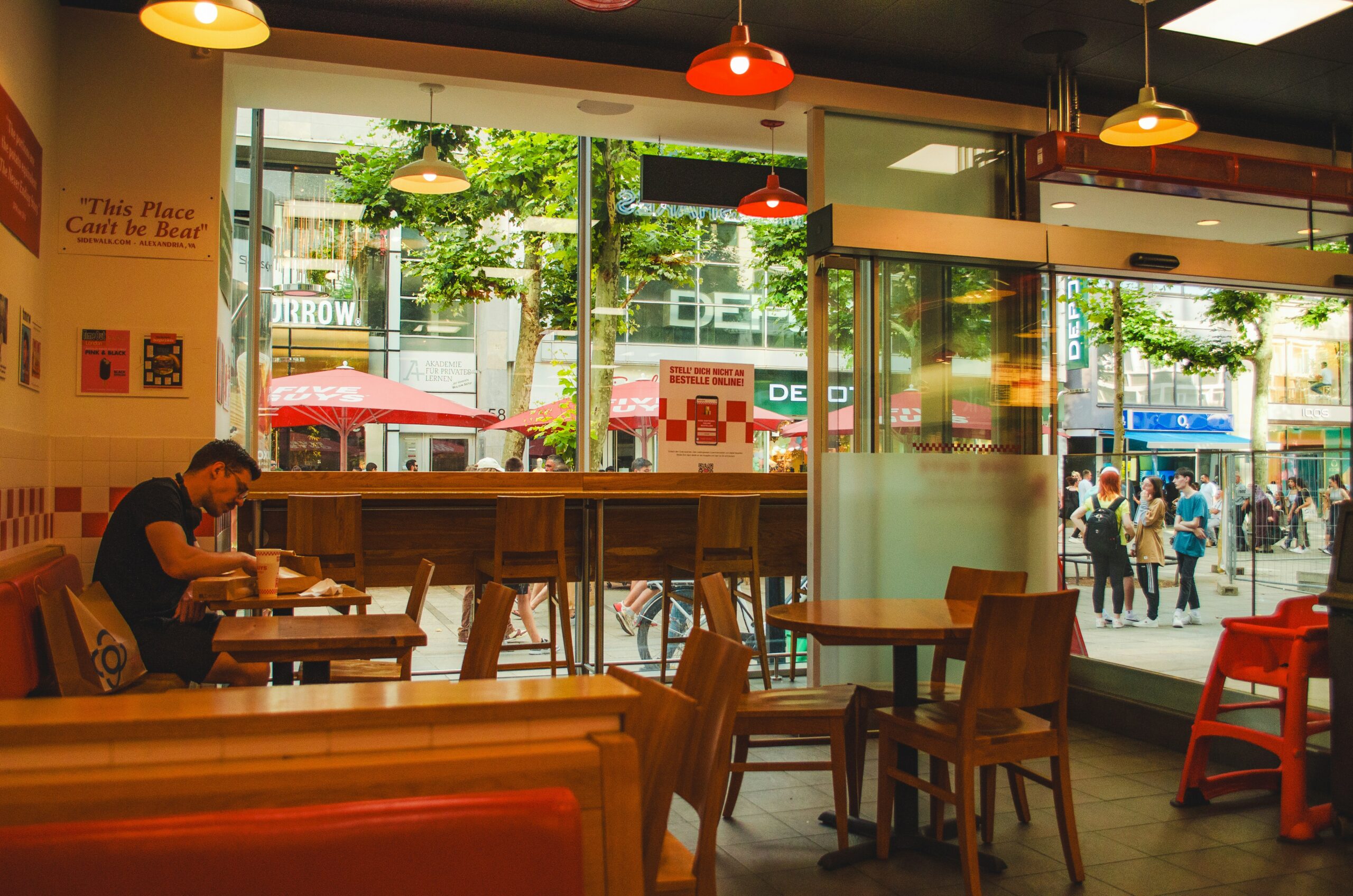 La photo montre l’intérieur d'un restaurant avec un bar en bois, des tabourets colorés et deux clients attablés au fond.