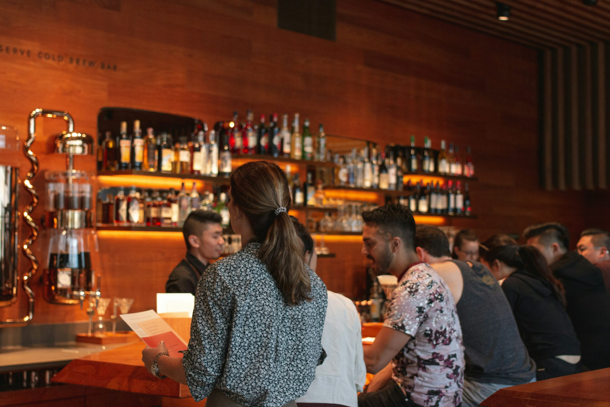 La photo montre une femme, tenant une carte de restaurant, face à un bar en bois où des clients sont assis. Un barman est en train de servir les clients.