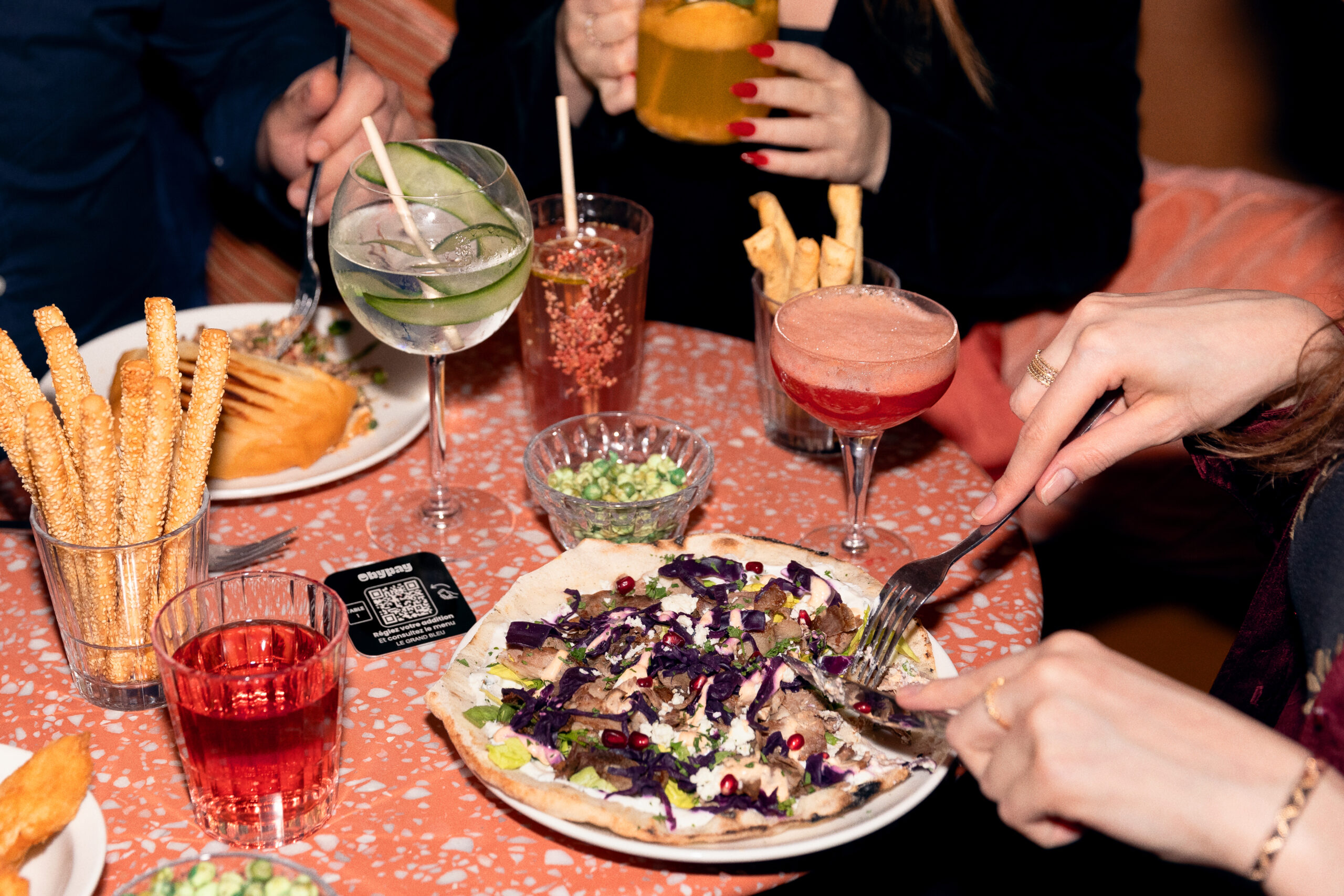 La photo montre un groupe de personnes partageant un repas dans un restaurant. Au centre, un QR Code est posé sur la table, permettant de payer l’addition.