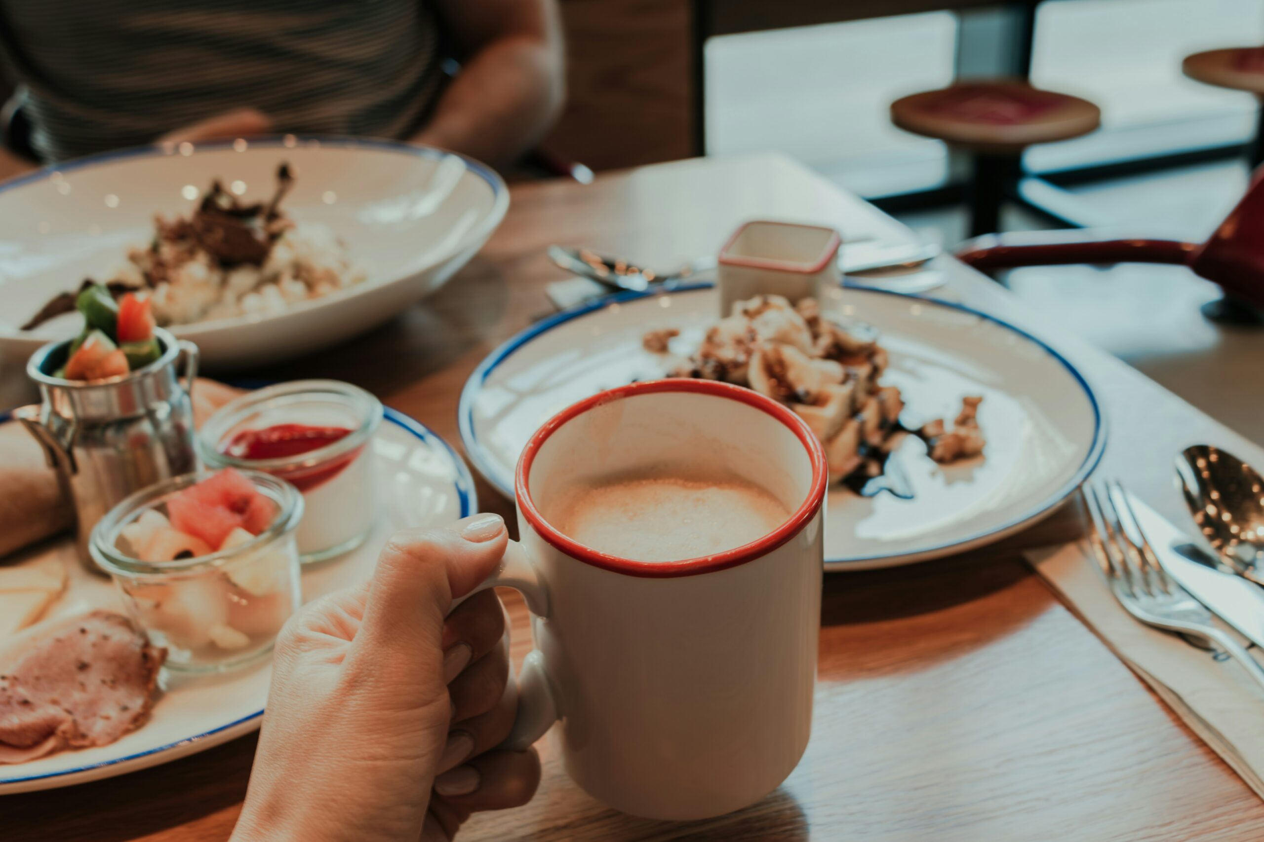 La photo montre une main tenant une tasse de café, posée sur une table avec un petit-déjeuner/brunch.