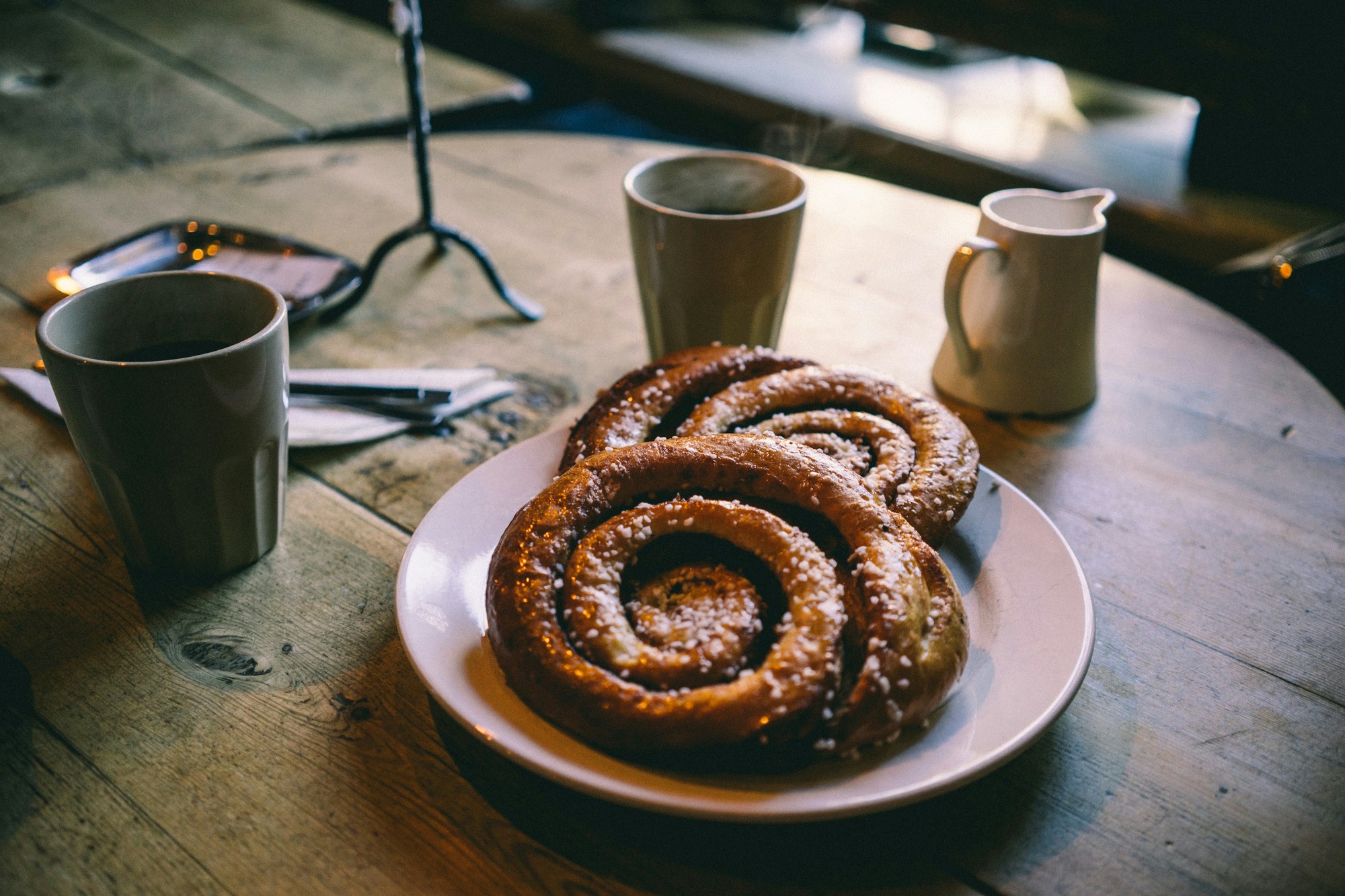 La photo montre une table en bois avec une assiette contenant deux viennoiseries, une tasse de café, une cruche et des couverts.