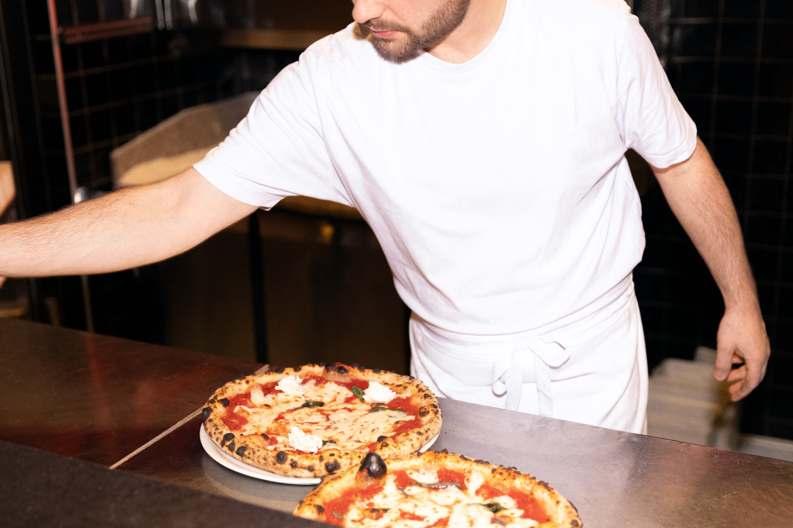 La photo montre un pizzaiolo en train de préparer des pizzas sur un comptoir de cuisine, dans un restaurant.