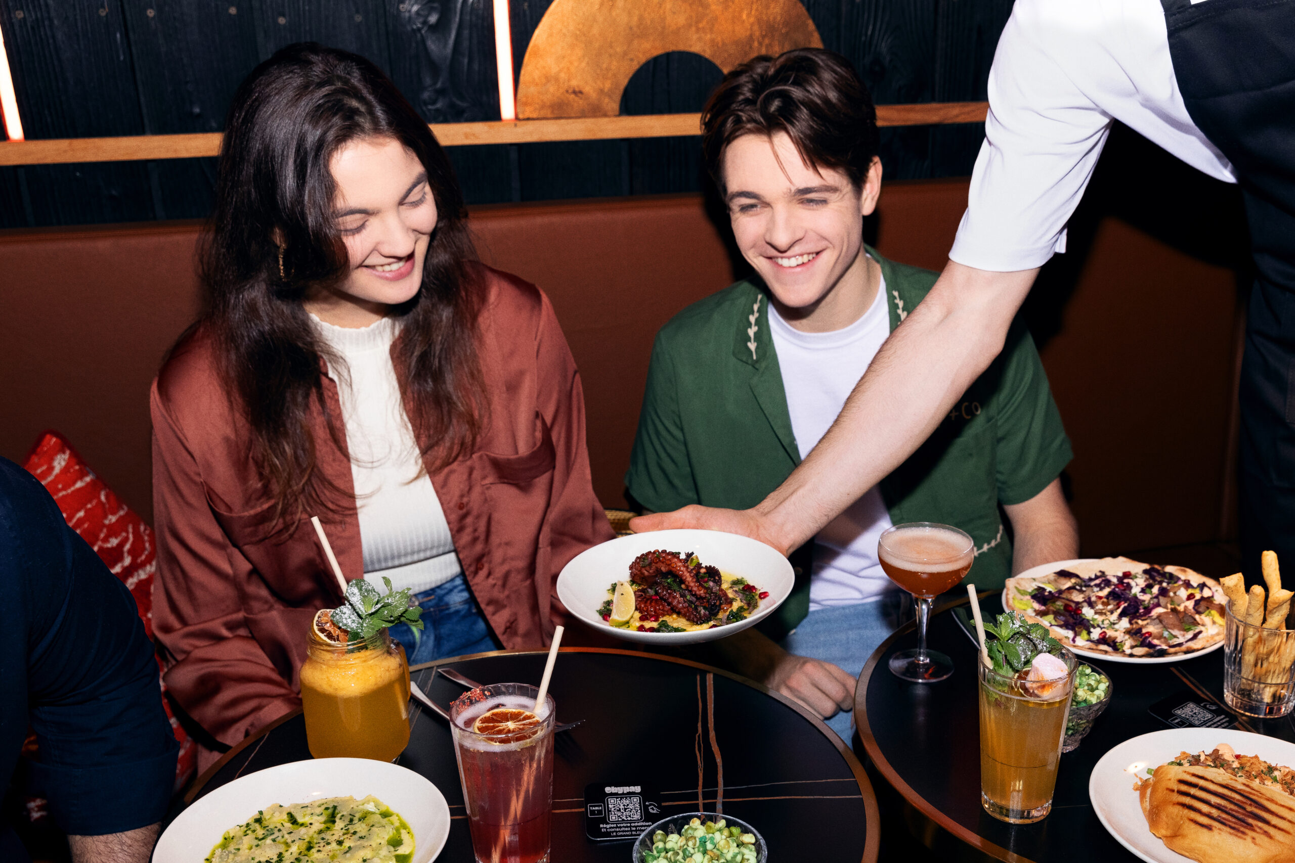 La photo montre un couple souriant dans un restaurant alors qu’un serveur leur apporte un plat. La table est remplie de cocktails et de plats variés. Un QR Code est visible sur la table, indiquant l’utilisation d’un système de commande digitale.