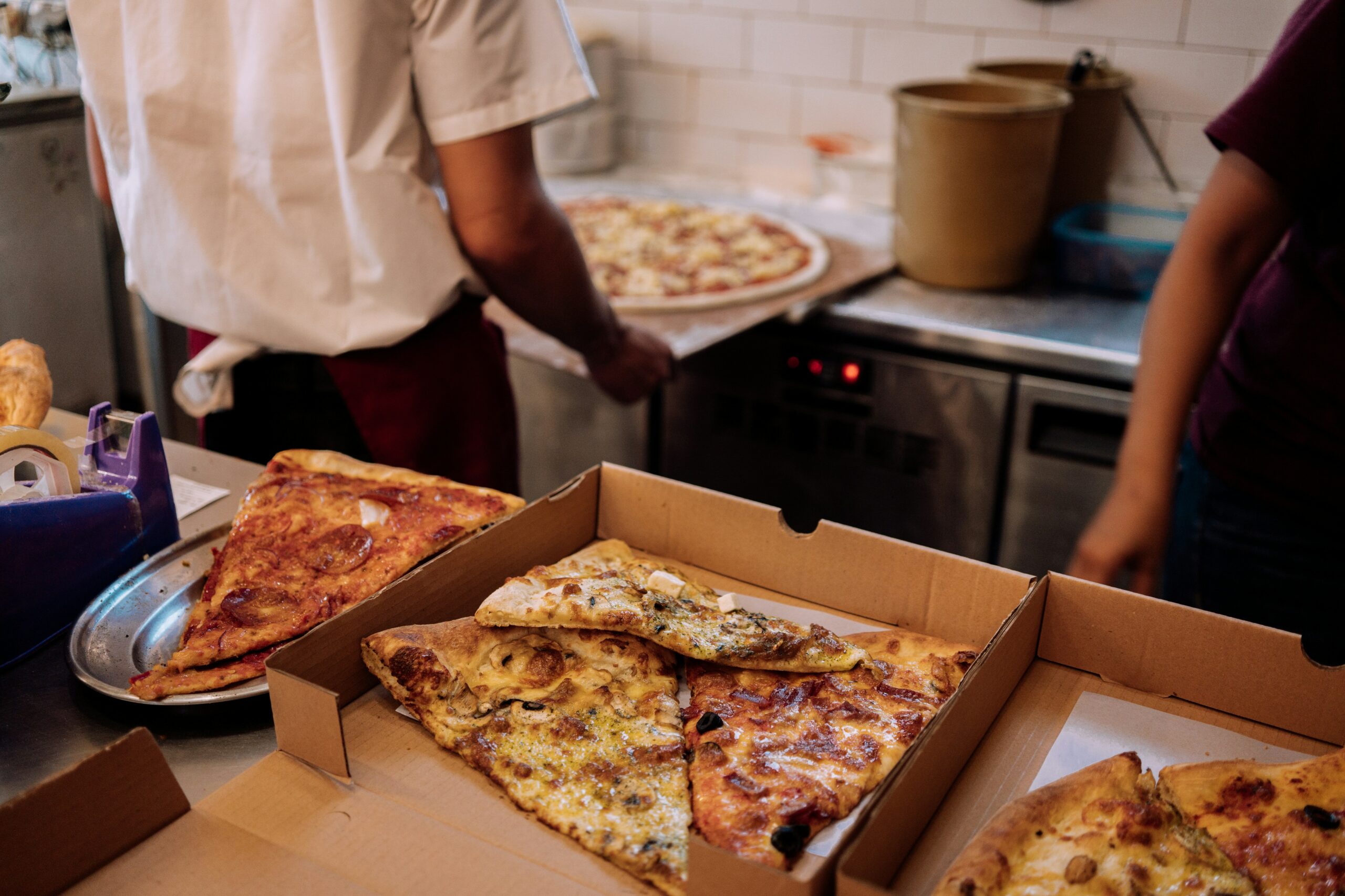 La photo montre une cuisine de restaurant. Plusieurs parts de pizza sont disposées dans des boîtes en carton prêtes pour une récupération en Click & Collect.