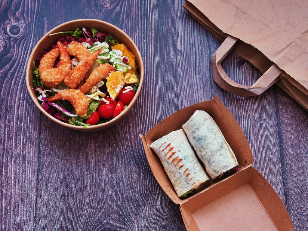 La photo montre une salade avec des crevettes panées, accompagnée de wraps dans une boîte en carton, posée sur une table en bois avec un sac kraft à côté.