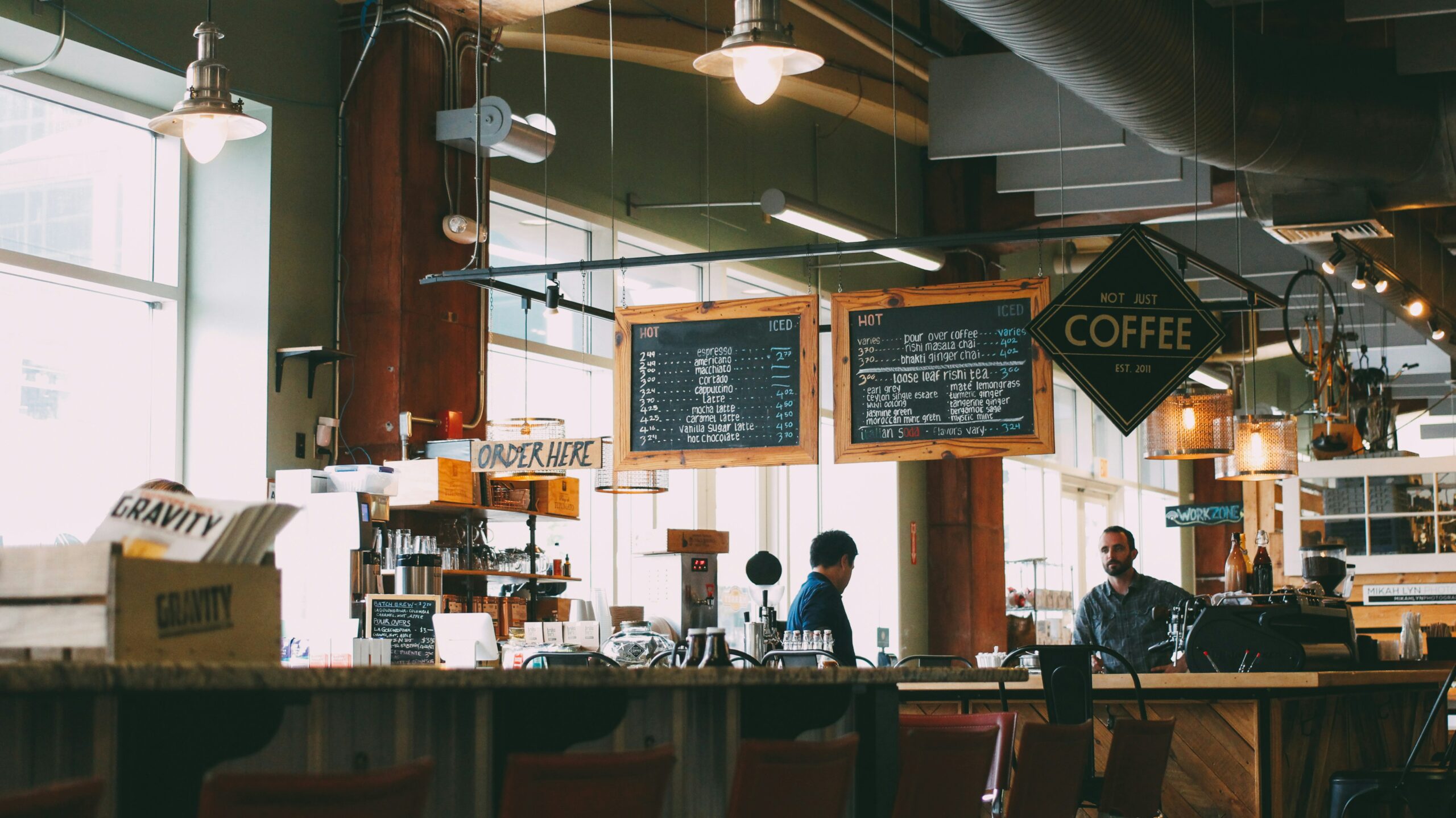 La photo montre un café avec un comptoir en bois, des tableaux de menu suspendus et une ambiance industrielle avec des luminaires suspendus. Deux employés sont visibles derrière le comptoir, tandis que des clients attendent ou passent commande.